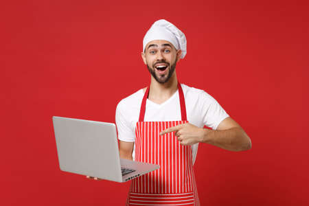 Excited young bearded chef cook or baker man in striped apron t-shirt toque chefs hat isolated on red background. Cooking food concept. Mock up copy space. Pointing index finger on laptop pc computer.の写真素材