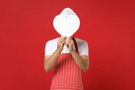 Young male chef cook or baker man in striped apron t-shirt toque chefs hat isolated on red background in studio. Cooking food concept. Mock up copy space. Covering face with white empty blank heart.の写真素材