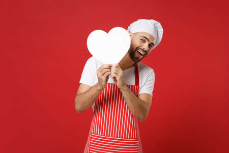 Funny young bearded male chef cook or baker man in striped apron t-shirt toque chefs hat isolated on red background. Cooking food concept. Mock up copy space. Holding in hands white empty blank heart.の写真素材