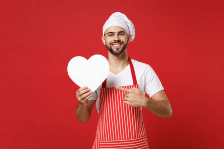 Smiling young bearded chef cook or baker man in striped apron toque chefs hat isolated on red background. Cooking food concept. Mock up copy space. Pointing index finger on white empty blank heart.の写真素材