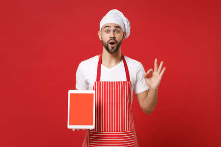 Shocked chef cook or baker man in striped apron toque chefs hat isolated on red background. Cooking food concept. Mock up copy space. Hold tablet computer with blank empty screen, showing OK gesture.の写真素材