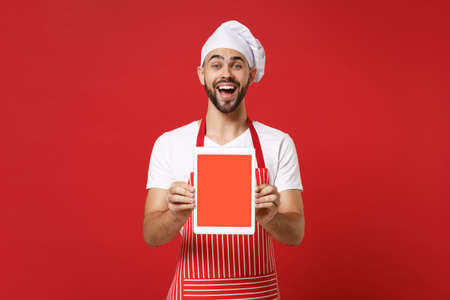 Excited young male chef cook or baker man in striped apron toque chefs hat isolated on red background. Cooking food concept. Mock up copy space. Holding tablet pc computer with blank empty screen.の写真素材