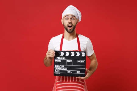 Shocked male chef cook or baker man in striped apron white t-shirt toque chefs hat isolated on red background. Cooking food concept. Mock up copy space. Holding classic black film making clapperboard.の写真素材