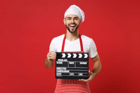 Cheerful male chef cook or baker man in striped apron white t-shirt toque chefs hat isolated on red background. Cooking food concept. Mock up copy space. Hold classic black film making clapperboard.の写真素材