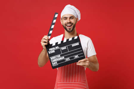 Laughing male chef cook or baker man in striped apron white t-shirt toque chefs hat isolated on red background. Cooking food concept. Mock up copy space. Hold classic black film making clapperboard.の写真素材