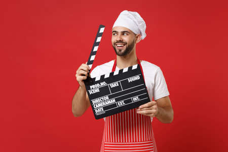 Funny male chef cook or baker man in striped apron white t-shirt toque chefs hat isolated on red background. Cooking food concept. Mock up copy space. Holding classic black film making clapperboard.の写真素材