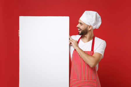 Joyful young bearded chef cook or baker man in striped apron white t-shirt toque chefs hat isolated on red wall background. Cooking food concept. Mock up copy space. Holding big empty blank billboard.の写真素材