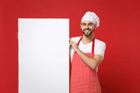 Smiling young bearded chef cook or baker man in striped apron white t-shirt toque chefs hat isolated on red background. Cooking food concept. Mock up copy space. Holding big empty blank billboard.の写真素材