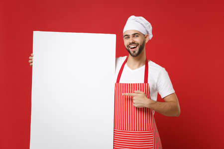 Cheerful chef cook or baker man in striped apron white t-shirt toque chefs hat isolated on red background. Cooking food concept. Mock up copy space. Pointing index finger on big empty blank billboard.の写真素材
