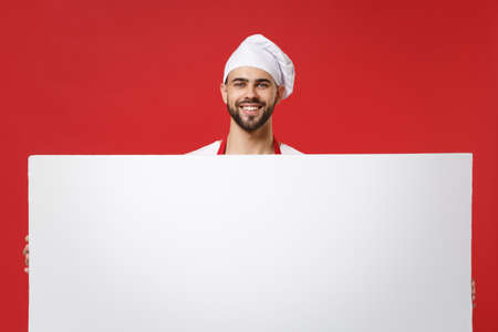 Smiling young male chef cook or baker man in toque chefs hat isolated on red background. Cooking food concept. Mock up copy space. Holding big white empty blank billboard with place for text or image.の写真素材