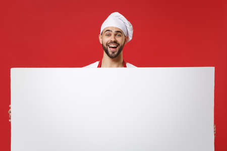 Cheerful young male chef cook or baker man in toque chefs hat isolated on red background. Cooking food concept. Mock up copy space. Hold big white empty blank billboard with place for text or image.の写真素材