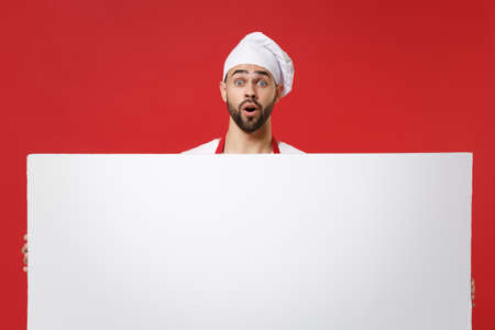 Shocked young male chef cook or baker man in toque chefs hat isolated on red background. Cooking food concept. Mock up copy space. Holding big white empty blank billboard with place for text or image.の写真素材