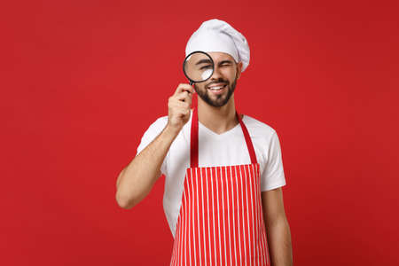 Smiling young bearded male chef cook or baker man in striped apron white t-shirt toque chefs hat isolated on red wall background. Cooking food concept. Mock up copy space. Holding magnifying glass.の写真素材