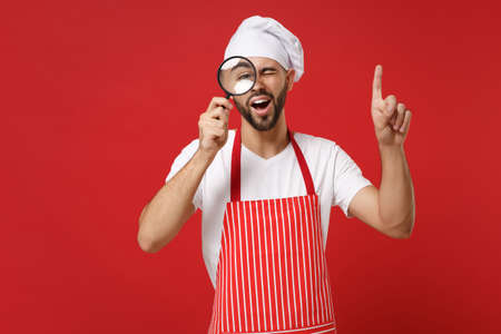 Chef cook or baker man in striped apron white t-shirt toque chefs hat isolated on red background. Cooking food concept. Mock up copy space. Hold magnifying glass, point index finger up with new idea.の写真素材