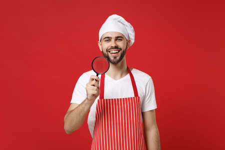 Smiling young bearded male chef cook or baker man in striped apron white t-shirt toque chefs hat isolated on red wall background. Cooking food concept. Mock up copy space. Holding magnifying glass.の写真素材