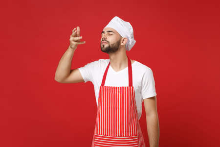 Young bearded male chef cook or baker man in striped apron white t-shirt toque chefs hat isolated on red background. Cooking food concept. Mock up copy space. Raised hand to face, feeling food smell.の写真素材
