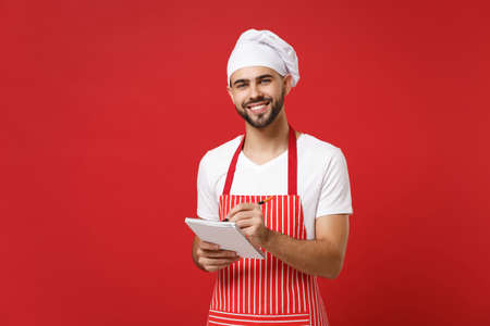 Smiling young bearded male chef cook or baker man in striped apron white t-shirt toque chefs hat isolated on red background. Cooking food concept. Mock up copy space. Holding notebook accepting order.の写真素材