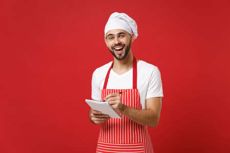 Cheerful young bearded male chef cook or baker man in striped apron white t-shirt toque chefs hat isolated on red background. Cooking food concept. Mock up copy space. Hold notebook, accepting order.の写真素材