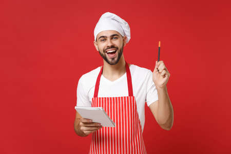 Cheerful young bearded male chef cook or baker man in striped apron white t-shirt toque chefs hat isolated on red background. Cooking food concept. Mock up copy space. Hold notebook, accepting order.の写真素材