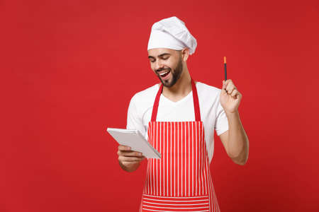 Cheerful young bearded male chef cook or baker man in striped apron white t-shirt toque chefs hat isolated on red background. Cooking food concept. Mock up copy space. Hold notebook, accepting order.の写真素材