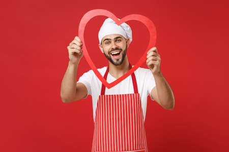 Cheerful young bearded male chef cook or baker man in striped apron white t-shirt toque chefs hat isolated on red wall background. Cooking food concept. Mock up copy space. Holding big wooden heart.の写真素材