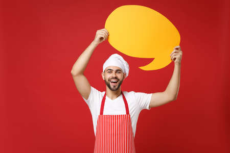 Laughing chef cook or baker man in striped apron white t-shirt toque chefs hat isolated on red background. Cooking food concept. Mock up copy space. Holding empty blank Say cloud yellow speech bubble.の写真素材