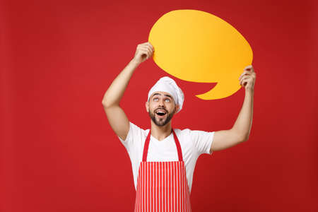 Amazed chef cook or baker man in striped apron white t-shirt toque chefs hat isolated on red background. Cooking food concept. Mock up copy space. Holding empty blank Say cloud, yellow speech bubble.の写真素材