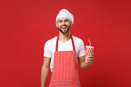 Cheerful young bearded male chef cook or baker man in striped apron white t-shirt toque chefs hat isolated on red background. Cooking food concept. Mock up copy space. Holding cup of cola or soda.の写真素材
