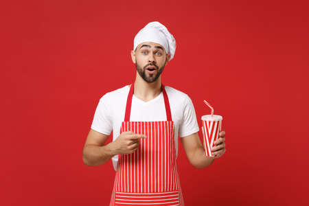 Shocked male chef cook or baker man in striped apron white t-shirt toque chefs hat isolated on red background. Cooking food concept. Mock up copy space. Pointing index finger on cup of cola or soda.の写真素材