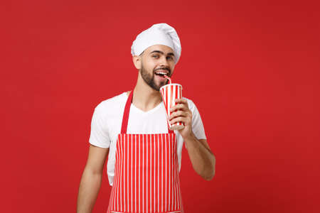 Handsome young bearded male chef cook or baker man in striped apron white t-shirt toque chefs hat isolated on red background. Cooking food concept. Mock up copy space. Holding cup of cola or soda.の写真素材