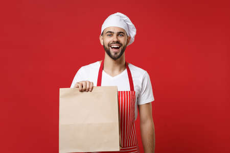 Excited male chef cook or baker man in striped apron toque chefs hat isolated on red background. Cooking food concept. Mock up copy space. Holding brown clear empty blank craft paper bag for takeaway.の写真素材