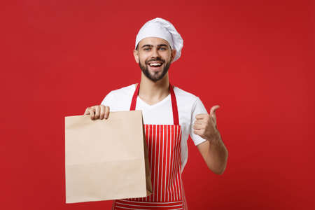 Chef cook baker man in striped apron toque chefs hat isolated on red background. Cooking food concept. Mock up copy space. Hold brown clear empty blank craft paper bag for takeaway, showing thumb up.の写真素材