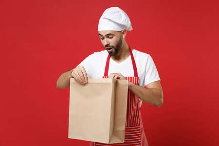 Curious male chef cook or baker man in striped apron toque chefs hat isolated on red background. Cooking food concept. Mock up copy space. Holding brown clear empty blank craft paper bag for takeaway.の写真素材