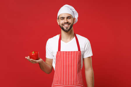 Smiling young male chef cook or baker man in striped apron white t-shirt toque chefs hat isolated on red wall background. Cooking food concept. Mock up copy space. Holding fresh vegetable bell pepper.の写真素材