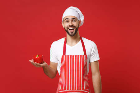 Excited young male chef cook or baker man in striped apron white t-shirt toque chefs hat isolated on red wall background. Cooking food concept. Mock up copy space. Holding fresh vegetable bell pepper.の写真素材