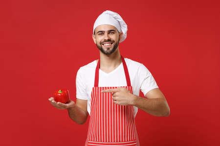 Smiling male chef cook or baker man in striped apron t-shirt toque chefs hat isolated on red background. Cooking food concept. Mock up copy space. Pointing index finger on fresh vegetable bell pepper.の写真素材