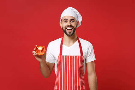 Joyful young male chef cook or baker man in striped apron white t-shirt toque chefs hat isolated on red background. Cooking food concept. Mock up copy space. Hold fresh vegetable half of bell pepper.の写真素材