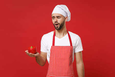 Perplexed young male chef cook or baker man in striped apron white t-shirt toque chefs hat isolated on red background. Cooking food concept. Mock up copy space. Holding fresh vegetable bell pepper.の写真素材