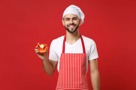 Smiling young male chef cook or baker man in striped apron white t-shirt toque chefs hat isolated on red background. Cooking food concept. Mock up copy space. Hold fresh vegetable half of bell pepper.の写真素材