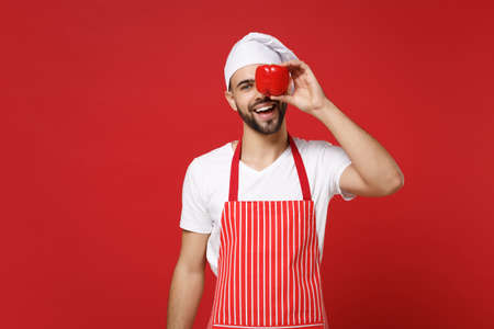 Cheerful male chef cook or baker man in striped apron white t-shirt toque chefs hat isolated on red background. Cooking food concept. Mock up copy space. Covering eye with fresh vegetable bell pepper.の写真素材