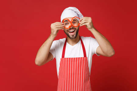 Funny male chef cook or baker man in striped apron toque chefs hat isolated on red background. Cooking food concept. Mock up copy space. Hold pepper slices near eyes, imitating glasses or binoculars.の写真素材