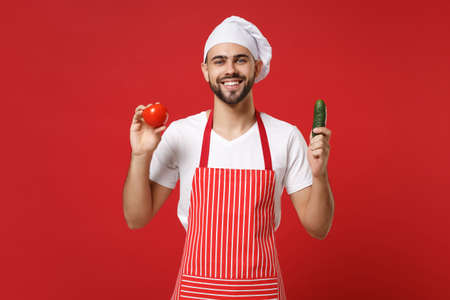 Smiling young male chef cook or baker man in striped apron white t-shirt toque chefs hat isolated on red background. Cooking food concept. Mock up copy space. Holding fresh vegetables tomato cucumber.の写真素材