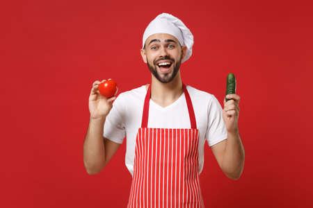 Excited young male chef cook or baker man in striped apron white t-shirt toque chefs hat isolated on red background. Cooking food concept. Mock up copy space. Holding fresh vegetables tomato cucumber.の写真素材