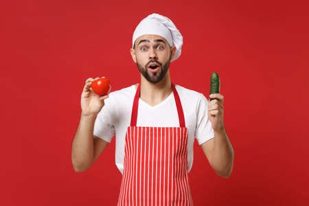 Shocked young male chef cook or baker man in striped apron white t-shirt toque chefs hat isolated on red background. Cooking food concept. Mock up copy space. Holding fresh vegetables tomato cucumber.の写真素材