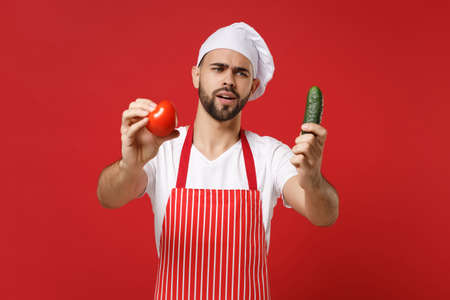 Puzzled young male chef cook or baker man in striped apron white t-shirt toque chefs hat isolated on red background. Cooking food concept. Mock up copy space. Holding fresh vegetables tomato cucumber.の写真素材