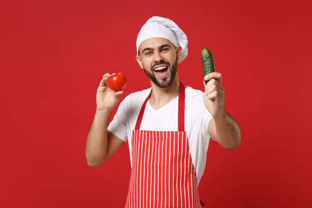 Funny young male chef cook or baker man in striped apron white t-shirt toque chefs hat isolated on red background. Cooking food concept. Mock up copy space. Holding fresh vegetables tomato, cucumber.の写真素材