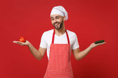 Young bearded male chef cook or baker man in striped apron white t-shirt toque chefs hat isolated on red background. Cooking food concept. Mock up copy space. Holding fresh vegetables tomato cucumber.の写真素材