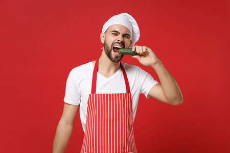 Young bearded male chef cook or baker man in striped apron white t-shirt toque chefs hat isolated on red background. Cooking food concept. Mock up copy space. Holding fresh vegetable, biting cucumber.の写真素材