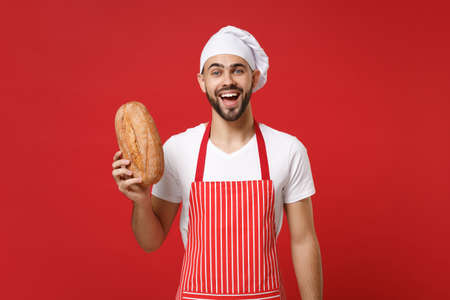 Excited young bearded male chef cook or baker man in striped apron white t-shirt toque chefs hat isolated on red background in studio. Cooking food concept. Mock up copy space. Holding bread in hand.の写真素材