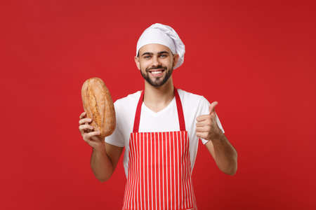 Smiling young bearded male chef cook or baker man in striped apron white t-shirt toque chefs hat isolated on red background. Cooking food concept. Mock up copy space. Holding bread, showing thumb up.の写真素材